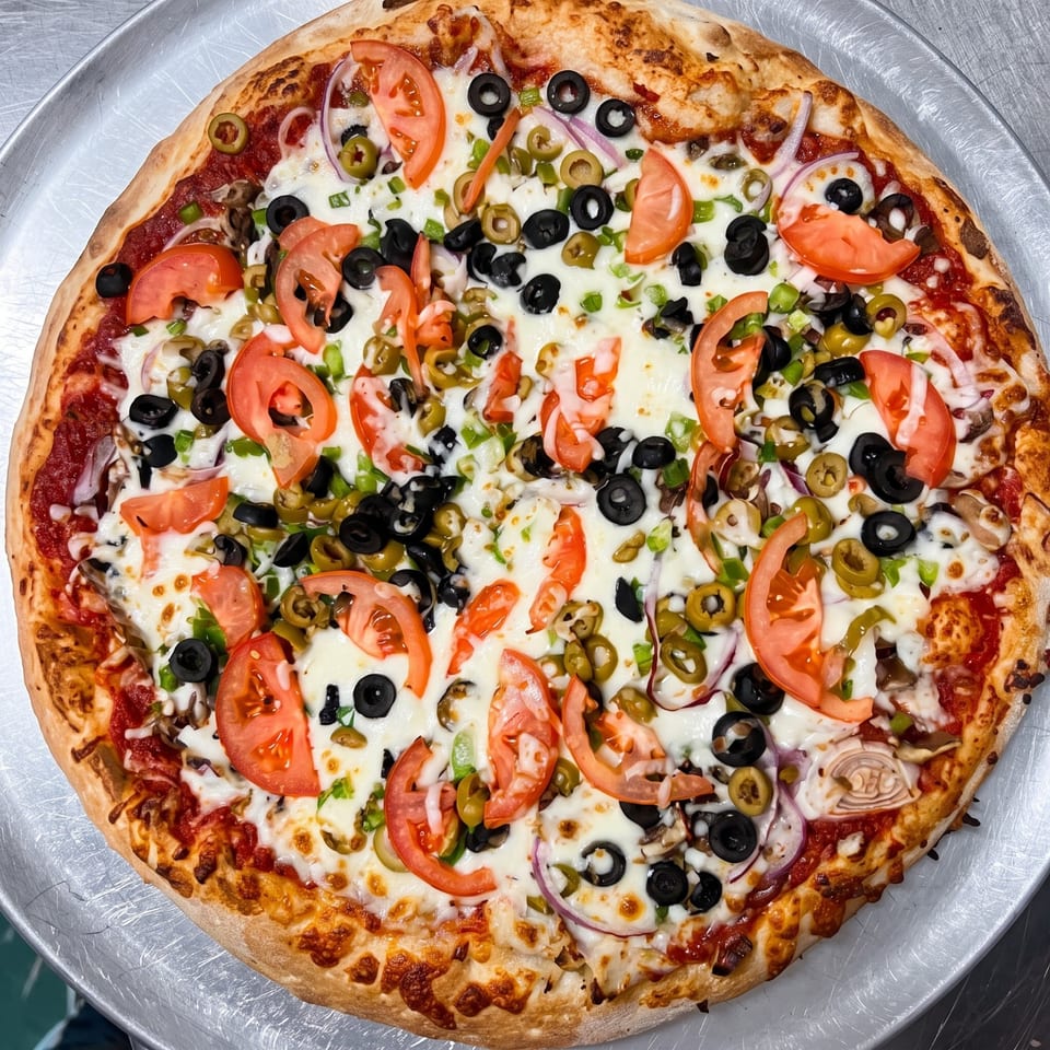 Large cheese pizza topped with tomatoes, black and green olives, jalapeños, green peppers, onions, and mushrooms on a metal tray.